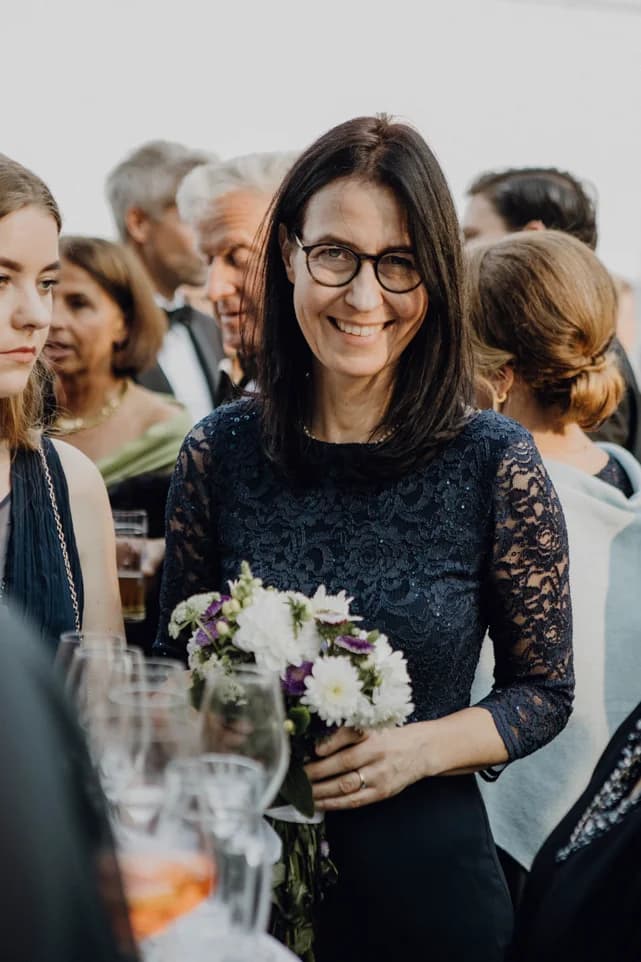 Beatrix „Trixi“ Diedenhofen lächelt mit einem Blumenstrauß in der Hand inmitten von Gästen bei einem Fest